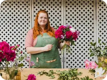 A person with long red hair in a green apron holds a bouquet of flowers