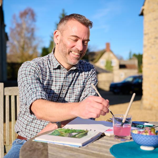 Person sits at an outdoor table, smiling while painting in a sketchbook, with paintbrushes and water nearby.